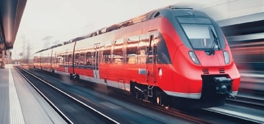 A modern red passenger train travels quickly along tracks at a station, with motion blur in the background indicating speed.