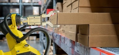 A yellow robotic arm with a gripper is picking up a cardboard box from a warehouse shelf filled with neatly stacked boxes.