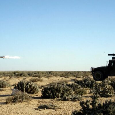 A military vehicle in a desert landscape launches a missile, which is seen mid-air with a visible trail of smoke. The sky is clear and the terrain is covered with sparse shrubs.