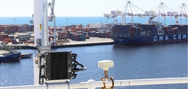 A communications device and antenna are mounted on a railing overlooking a busy seaport with stacked shipping containers and a large cargo ship docked near cranes by the water.