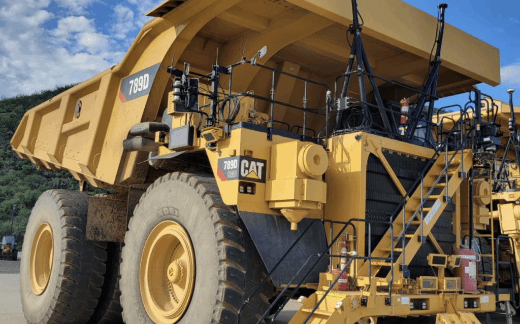 A large yellow Caterpillar 789D mining dump truck is parked outdoors on a sunny day, with metal stairs leading to the cab and hills visible in the background.