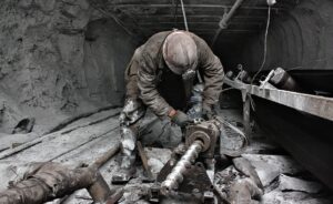 A miner wearing a helmet and headlamp operates machinery in a dark, dusty underground tunnel, surrounded by scattered industrial equipment and covered in gray dust.