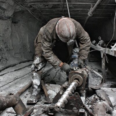 A miner wearing a helmet and headlamp operates machinery in a dark, dusty underground tunnel, surrounded by scattered industrial equipment and covered in gray dust.
