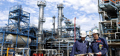 Two workers in safety gear stand in front of an industrial oil refinery or chemical plant with metal structures, storage tanks, and tall pipes against a blue sky.