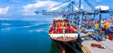 A large cargo ship loaded with colorful shipping containers is docked at a port, with cranes unloading containers beside calm blue water and a clear sky.