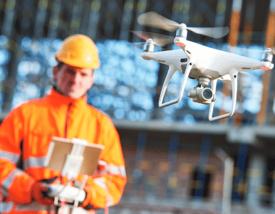 A person in an orange safety jacket and hard hat operates a drone using a remote control at a construction site. The drone is flying in the foreground with a blurred structure in the background.