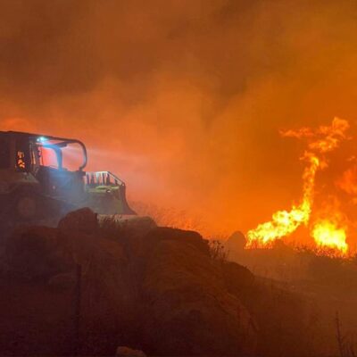 A bulldozer with headlights on is seen at night near intense, tall flames and heavy smoke from a wildfire, as it moves along a dirt path surrounded by burning vegetation.
