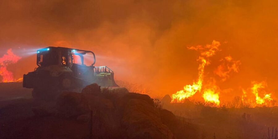 A bulldozer with headlights on is seen at night near intense, tall flames and heavy smoke from a wildfire, as it moves along a dirt path surrounded by burning vegetation.