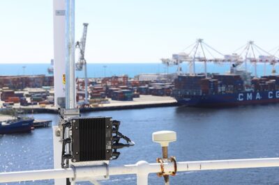 A close-up of a maritime communication device mounted on a railing overlooks a busy shipping port with stacked cargo containers, cranes, and a large container ship docked by the water.