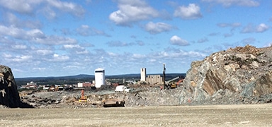 A rocky open landscape with construction equipment at the center, surrounded by large rock piles. In the distance, there are industrial buildings and a partly cloudy blue sky.