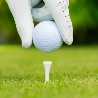 A gloved hand holds a white golf ball above a white tee on green grass, preparing to place the ball for a shot. The background is blurred with hints of greenery.