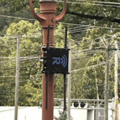 A black wireless communication device with an antenna is mounted on a tall orange utility pole, surrounded by numerous power lines and green trees in the background.