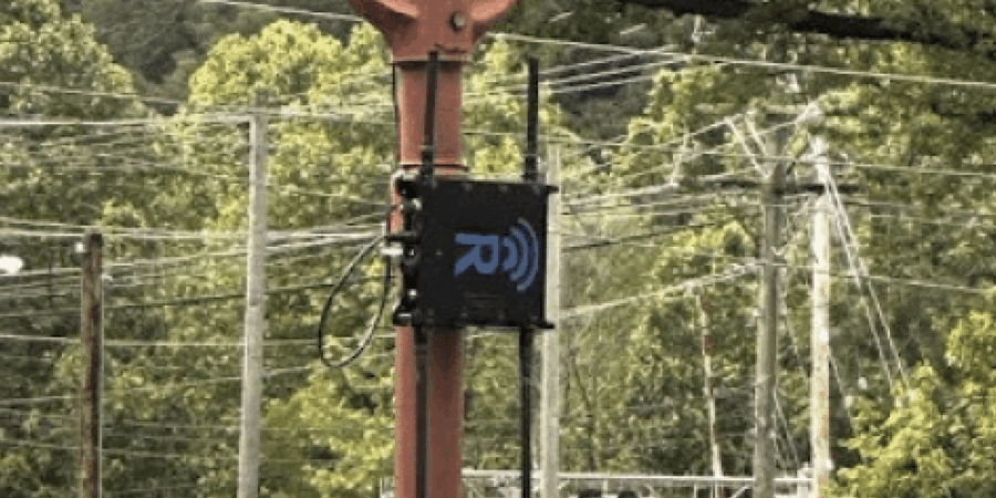 A black wireless communication device with an antenna is mounted on a tall orange utility pole, surrounded by numerous power lines and green trees in the background.