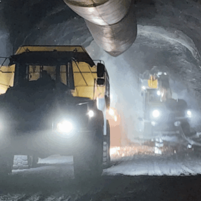 Two large mining trucks with headlights on drive through a dimly lit, dusty underground tunnel with a ventilation pipe overhead.