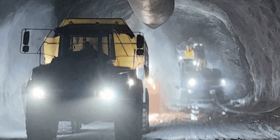 Two large mining trucks with headlights on drive through a dimly lit, dusty underground tunnel with a ventilation pipe overhead.