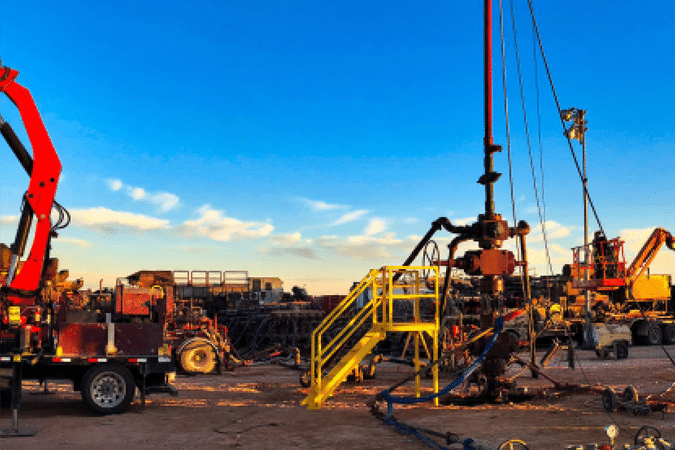 Oil drilling rig site at sunset with machinery, pipelines, and equipment; a red crane and a yellow staircase are visible, with workers and vehicles scattered across the area under a clear blue sky.