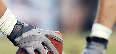 A football player wearing gloves prepares to snap a football, with another player's legs visible in the background.