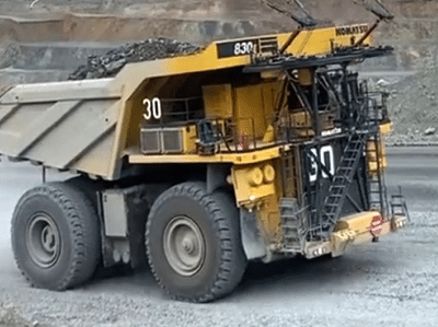 A large yellow mining dump truck, numbered 30, drives along a gravel road at an open-pit mine, carrying a load of rocks in its rear bed.