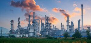 An industrial refinery complex with tall chimneys and metal structures emits smoke into the sky at sunset, with green vegetation in the foreground.