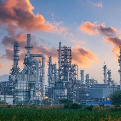 An industrial refinery complex with tall chimneys and metal structures emits smoke into the sky at sunset, with green vegetation in the foreground.