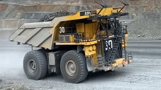 A large yellow mining dump truck, numbered 30, drives along a gravel road at an open-pit mine, carrying a load of rocks in its rear bed.