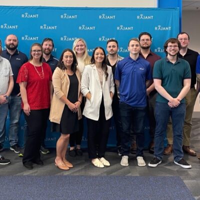 A group of fifteen people, men and women, stand and smile in front of a blue backdrop with the Rajant logo in an office setting. The group is casually dressed and posing for a team photo.