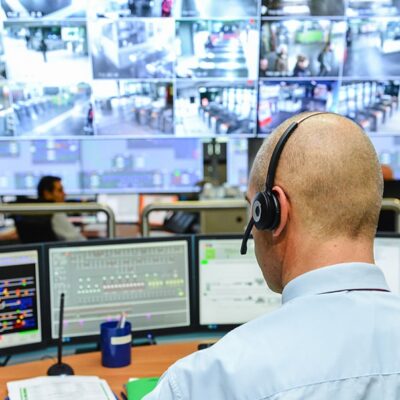A person wearing a headset monitors multiple computer screens and surveillance camera feeds in a control room, with another person seated in the background, also working.