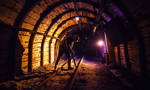 A person wearing a helmet and headlamp walks inside a dimly lit, wooden-supported mining tunnel, with tracks running along the ground and purple lights illuminating parts of the tunnel.