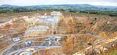 A large open-pit mine with terraced walls, exposed rock layers, and mining equipment at the bottom, set against a backdrop of green hills and a cloudy sky.