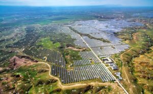 Aerial view of a vast solar farm with hundreds of solar panels spread across a green landscape, intersected by dirt roads and buildings, under a clear sky with faint digital network overlay elements.