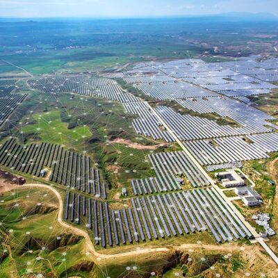 Aerial view of a vast solar farm with hundreds of solar panels spread across a green landscape, intersected by dirt roads and buildings, under a clear sky with faint digital network overlay elements.