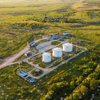Aerial view of a rural industrial facility with large white storage tanks, surrounded by green fields, buildings, and roads, with digital network graphics overlaid on the image.