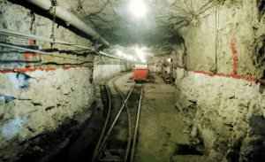 A dimly lit underground mine tunnel with rough stone walls, metal pipes, rail tracks, and a red mining cart at the center. Red markings line the tunnel walls, and equipment is scattered along the sides.
