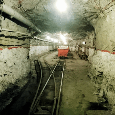 A dimly lit underground mine tunnel with rough stone walls, metal pipes, rail tracks, and a red mining cart at the center. Red markings line the tunnel walls, and equipment is scattered along the sides.