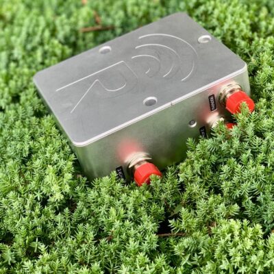 A metal electronic device with red connectors is partially surrounded by dense, green, bushy plants outdoors. The device has a logo with curved lines engraved on its top surface.
