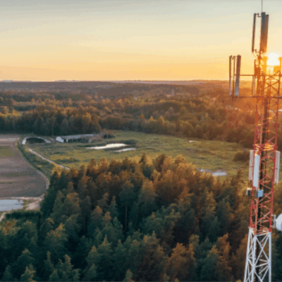 A tall red and white telecommunications tower stands amid a forest and fields at sunset, with the sun glowing in the background and a rural landscape stretching into the distance.