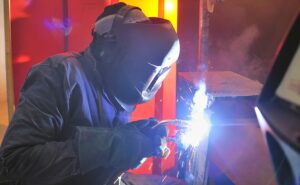 A person wearing protective gear, including a helmet and gloves, is welding metal indoors. Bright sparks and light are visible at the point of contact between the welding tool and the metal surface.