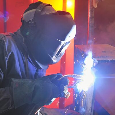 A person wearing protective gear, including a helmet and gloves, is welding metal indoors. Bright sparks and light are visible at the point of contact between the welding tool and the metal surface.