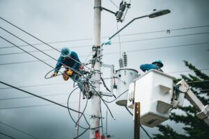 Two utility workers wearing safety gear repair electrical wires on a pole; one climbs the pole while the other operates from a bucket truck. The sky is overcast.