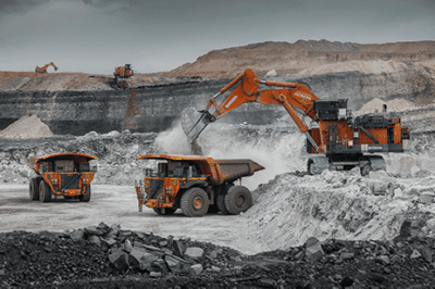 Large orange mining trucks and an excavator operate in an open-pit mine, surrounded by layers of earth and rock under a cloudy sky. Dust rises as machinery loads rocks into the trucks.