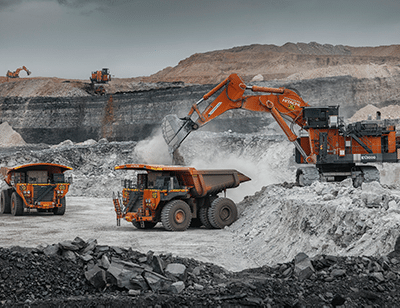Large orange mining trucks and an excavator operate in an open-pit mine, surrounded by layers of earth and rock under a cloudy sky. Dust rises as machinery loads rocks into the trucks.