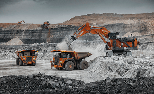 Large orange mining trucks and an excavator operate in an open-pit mine, surrounded by layers of earth and rock under a cloudy sky. Dust rises as machinery loads rocks into the trucks.