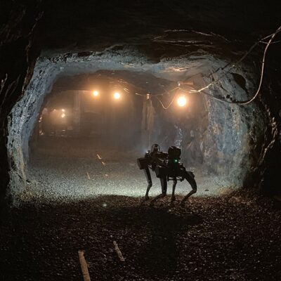A four-legged robot stands in a dimly lit, rocky underground tunnel with exposed wires and several bright lights illuminating the misty passage ahead.