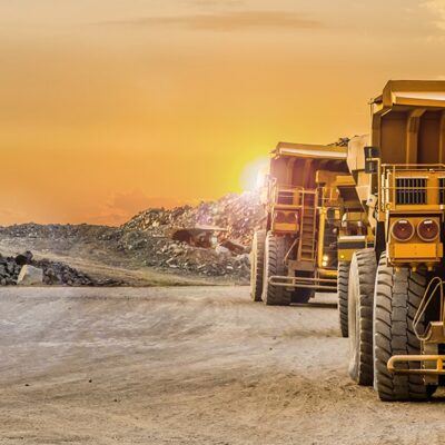 Two large yellow mining dump trucks drive on a dirt road at a mining site, surrounded by piles of rocks and gravel, with the sun setting in the background.