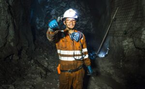 A miner wearing an orange jumpsuit, helmet with a headlamp, gloves, and safety gear stands underground in a dimly lit mine, giving a thumbs-up and smiling at the camera.