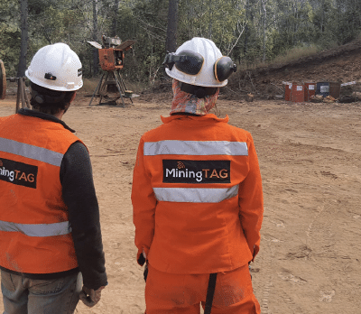 Two workers in orange safety gear and white helmets, with "MiningTAG" logos on their vests, stand on a dirt worksite surrounded by trees and equipment.