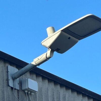 A modern streetlight is mounted on the side of a gray, corrugated building against a clear blue sky. Wires and a small electrical box are attached to the mounting pole.