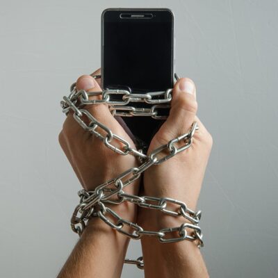 Two hands holding a smartphone are tightly wrapped in heavy metal chains, symbolizing addiction or being trapped by technology. The background is plain and light gray.