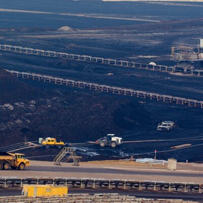 A large open-pit mining site with dark earth, several conveyor belts, heavy machinery, trucks, and small buildings scattered throughout the landscape.