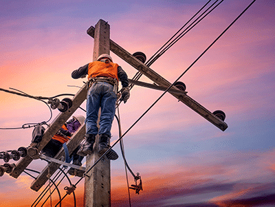 Two utility workers in orange vests and helmets repair power lines on a tall wooden pole at sunset, with a colorful sky in the background.
